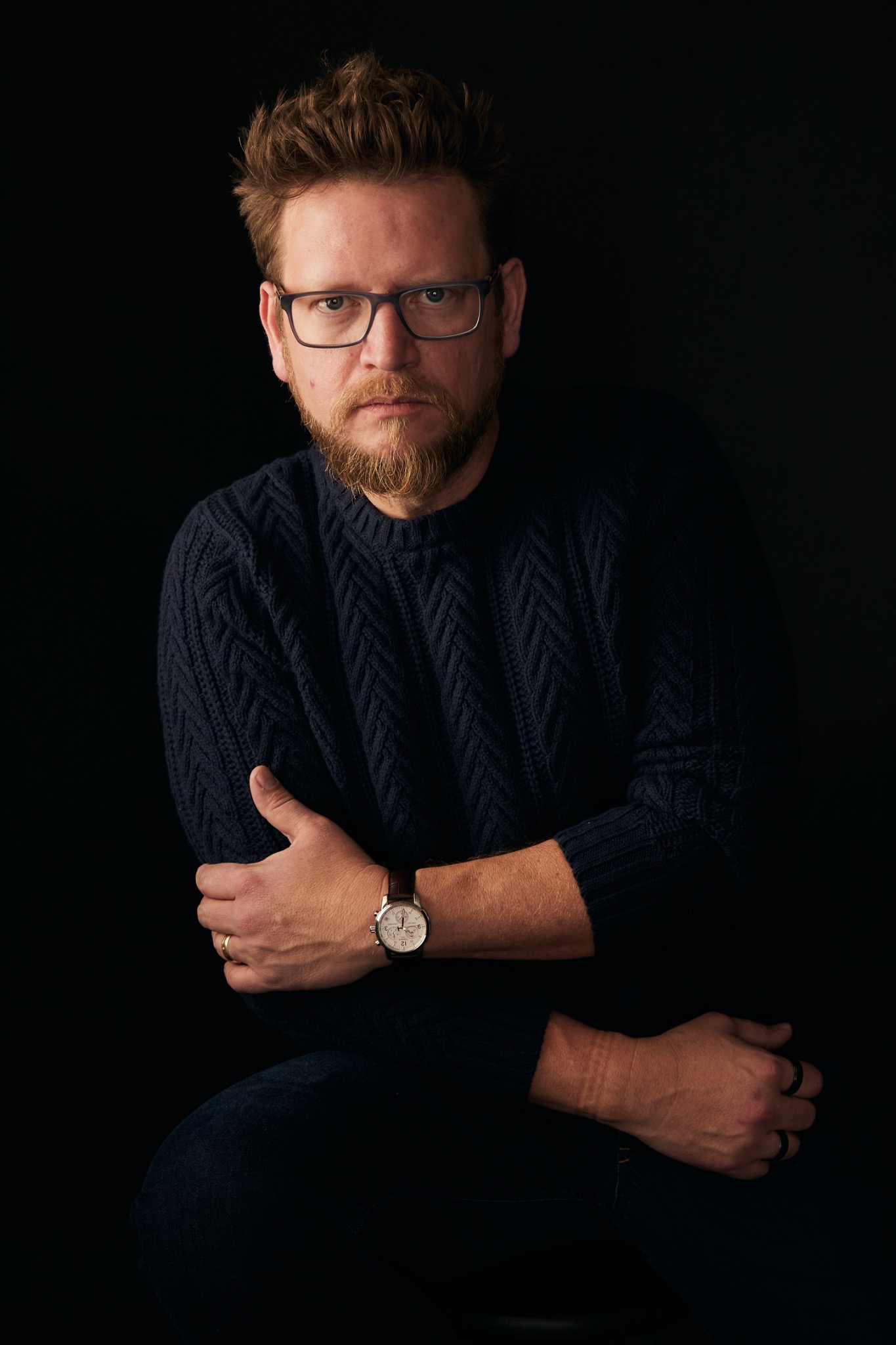 Aigo Pyles seated with his arms crossed, wearing a dark sweater and glasses.